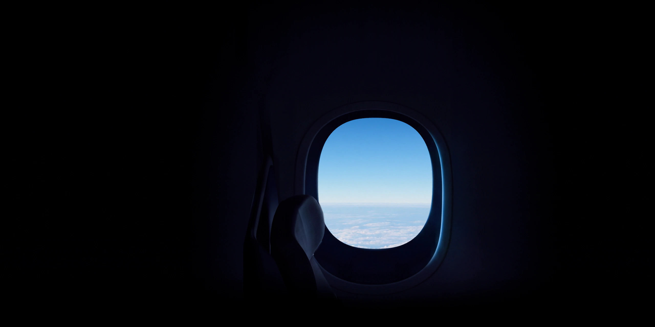 An aircraft window in a darkened cabin, beside a seat, looking out on a blue sky over a deck of cloud