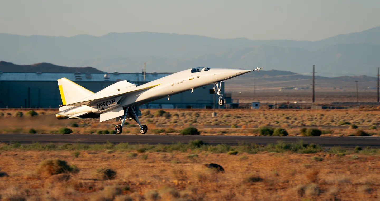 XB-1 lifting off the runway, its nose high, with a hangar and mountains behind